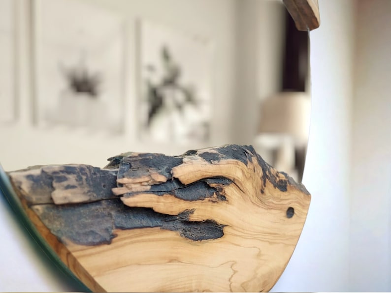 Puede incluir: Un espejo de pared circular con un marco de madera natural gruesa con borde vivo. La madera muestra una veta de color tostado claro con secciones oscuras, carbonizadas o negras desgastadas a lo largo de la superficie irregular.