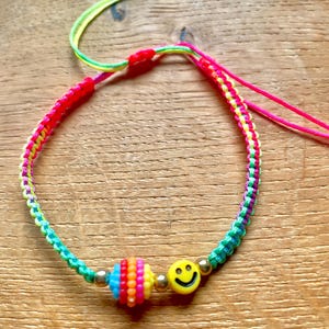 Peut inclure: Un bracelet en cordon tressé avec des fils néon rose, jaune, vert et violet. Le bracelet comprend une perle ronde à rayures multicolores, une perle jaune en forme de visage souriant et de petites perles intercalaires dorées métalliques.