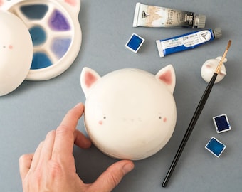 May include: A white ceramic cat-shaped box with pink ears and a painted face. The box is open and the inside is empty. There are paint tubes, paintbrushes, and watercolor paints in the background.