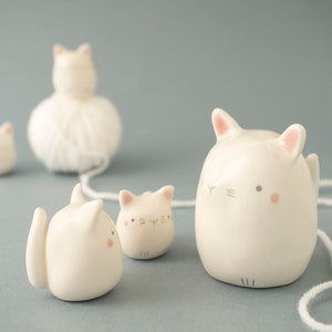 Puede incluir: Cinco figuritas de gato de cerámica blanca, una sosteniendo una bola de lana, con acentos rosados en las orejas y las mejillas. Los gatos están dispuestos en línea, con el gato de lana en el medio.