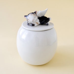 May include: White ceramic urn with a lid featuring a sleeping cat with wings. The cat is black, white, and brown.
