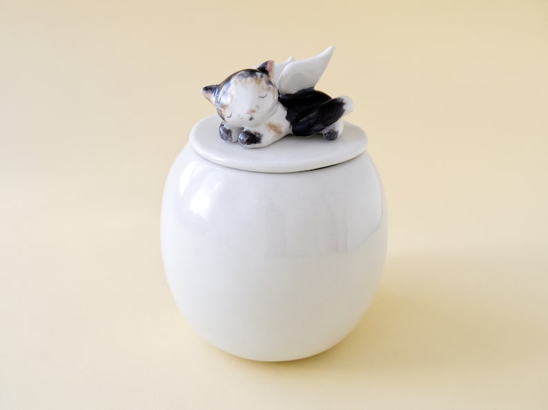 May include: White ceramic urn with a lid featuring a sleeping cat with wings. The cat is black, white, and brown.