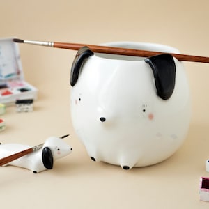 Könnte beinhalten: Zwei weiße, keramik-dachshundförmige Pinselhalter. Der größere Halter hat einen braunen Pinsel, der auf dem Rand liegt. Der kleinere Halter hat einen braunen Pinsel, der obenauf liegt. Beide Halter haben schwarze Ohren und gemalte Augen.