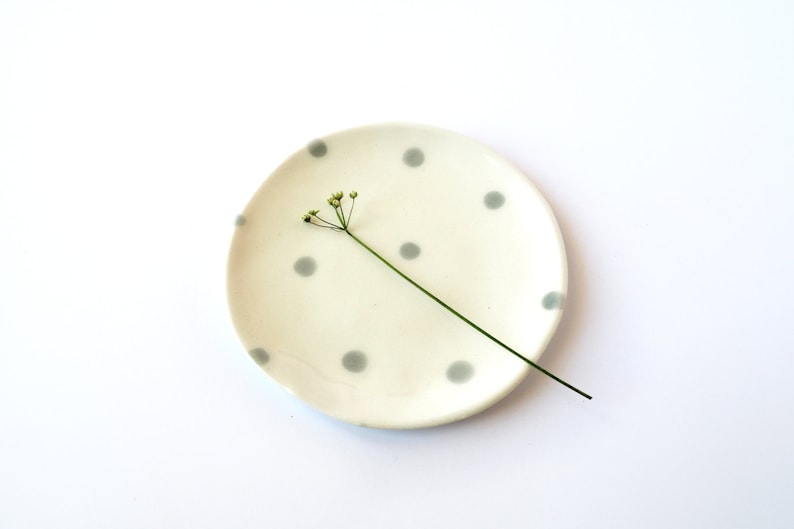 May include: A small, white ceramic dish with a grey polka dot pattern. A single sprig of green foliage sits on the dish.