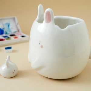 May include: Two white ceramic bunny figurines. The larger bunny has a pink nose and ears and is shaped like a cup. The smaller bunny is sitting upright.