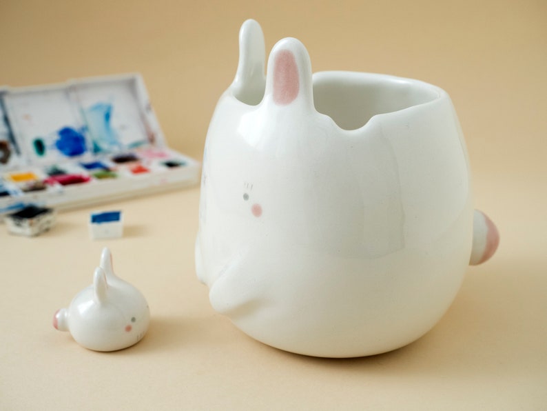 May include: Two white ceramic bunny figurines. The larger bunny has a pink nose and ears and is shaped like a cup. The smaller bunny is sitting upright.