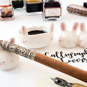 May include: White ceramic bunny inkwell with black ink, two white ceramic bunny pen holders, a wooden calligraphy pen with a silver tip, and a black ink bottle with a label that says "Siena Sophia" on a white surface with the words "Calligraphy Lovers" written in black ink.