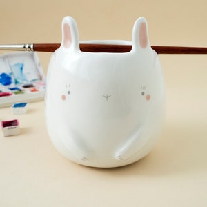 May include: White ceramic brush holder shaped like a bunny with pink ears and painted eyes. The holder is sitting on a light beige surface with a paintbrush resting on top.