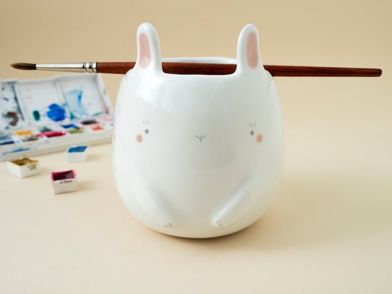 May include: White ceramic brush holder shaped like a bunny with pink ears and painted eyes. The holder is sitting on a light beige surface with a paintbrush resting on top.