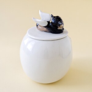 May include: White ceramic jar with a lid featuring a black cat with white wings. The cat is lying on its side with its head resting on the lid.