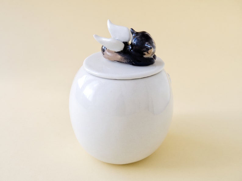 May include: White ceramic jar with a lid featuring a black cat with white wings. The cat is lying on its side with its head resting on the lid.