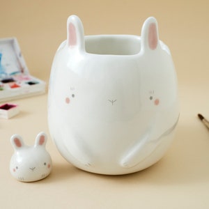 May include: Two white ceramic bunny figurines. The larger bunny has a wide, round body and pink ears. The smaller bunny is a smaller version of the larger bunny.