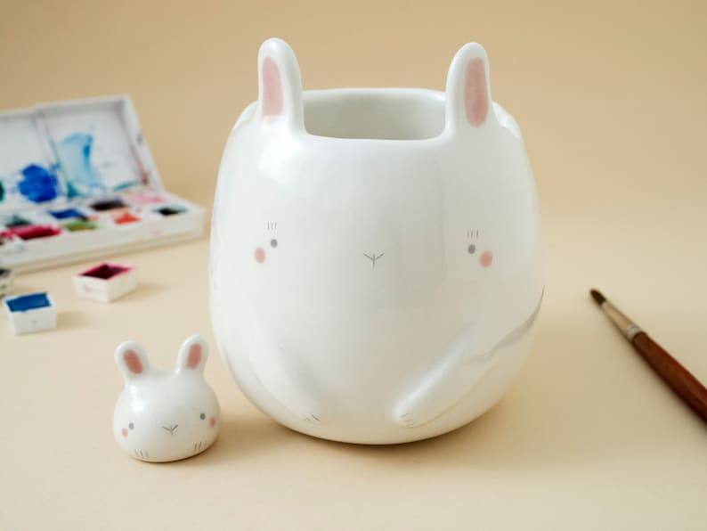 May include: Two white ceramic bunny figurines. The larger bunny has a wide, round body and pink ears. The smaller bunny is a smaller version of the larger bunny.