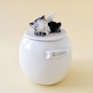 May include: White ceramic pet urn with a sleeping cat figurine on the lid. The cat has black and brown fur and white wings. The urn has the name "Romeo" written on it.