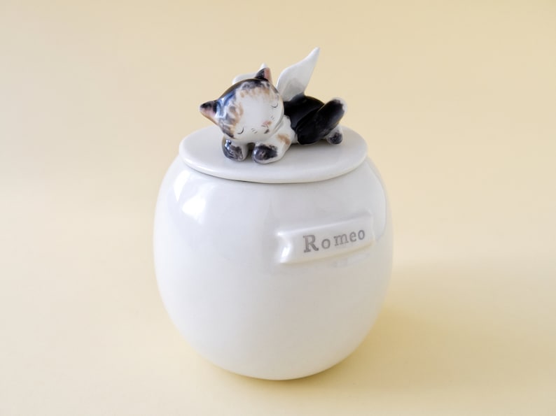 May include: White ceramic pet urn with a sleeping cat figurine on the lid. The cat has black and brown fur and white wings. The urn has the name "Romeo" written on it.
