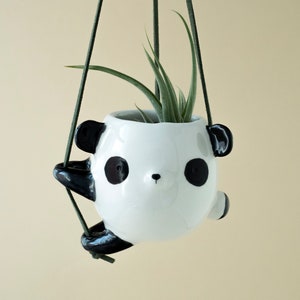 Puede incluir: Un macetero de cerámica blanca con forma de panda con orejas, ojos y nariz negros, colgado de cordones verdes. El macetero está lleno de una planta de aire verde.