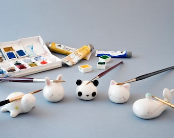 May include: Four ceramic animal brush holders, a panda, a rabbit, a cat, and a whale, with paintbrushes in each. The holders are white with painted details. There are also watercolor paints and paint tubes in the background.