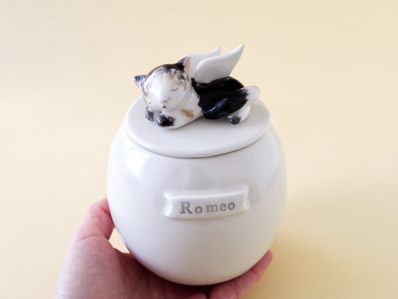 May include: White ceramic pet urn with a sleeping cat figurine on the lid. The cat has black, white, and brown fur and angel wings. The urn has the name "Romeo" inscribed on the front.