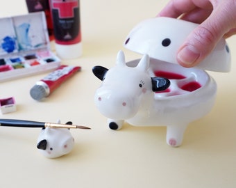 Puede incluir: Un recipiente de cerámica blanca con forma de vaca con una tapa extraíble. La tapa tiene manchas negras y la vaca tiene orejas y pezuñas negras. El recipiente tiene un interior rosa con tres compartimentos. Una vaca de cerámica blanca más pequeña con orejas y pezuñas negras está en primer plano. Un pincel descansa sobre la vaca más pequeña.