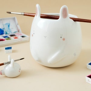 Könnte beinhalten: Weißer Keramik-Pinselhalter in Form eines Hasen mit rosa Ohren und einem darauf liegenden Pinsel. Im Vordergrund befindet sich ein kleinerer Hasen-Pinselhalter. Aquarellfarben sind im Hintergrund.