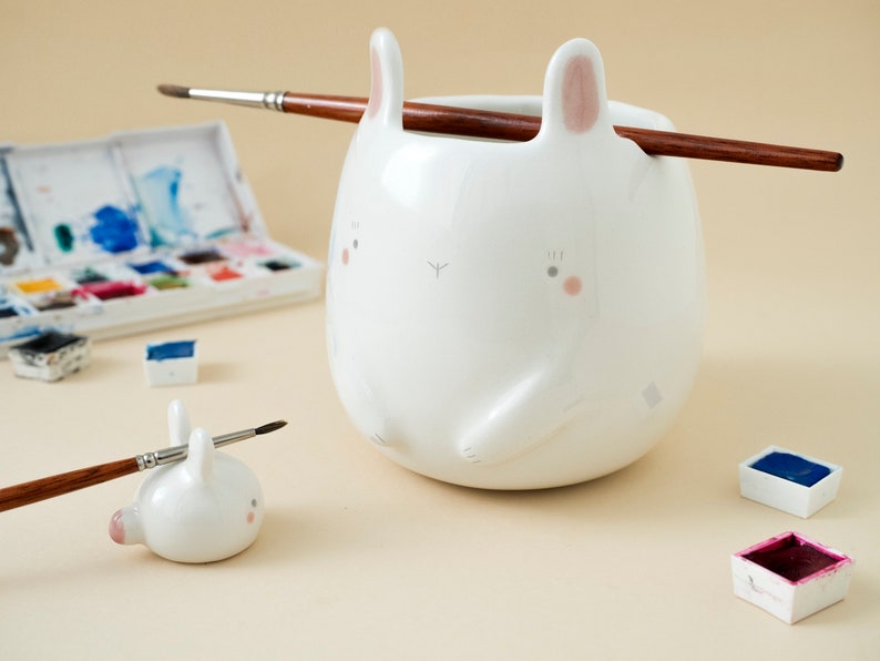 May include: White ceramic bunny brush holder with pink ears and a brush resting on top. A smaller bunny brush holder is in the foreground. Watercolour paints are in the background.