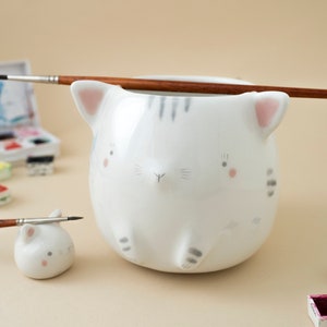 May include: Two white ceramic cat-shaped brush holders. The larger one has a grey stripe and pink ears. The smaller one has pink ears. Both have painted details on their faces. A paintbrush is resting on the larger holder.