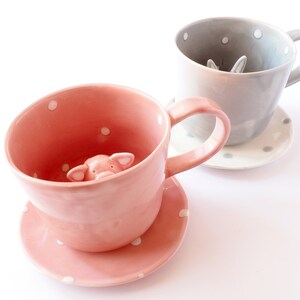 May include: Two ceramic teacups with matching saucers. The pink teacup has a small pig figurine peeking out of the cup. The grey teacup has a small rabbit figurine peeking out of the cup.