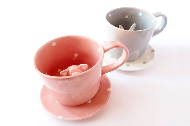 May include: Two ceramic teacups with matching saucers. The pink teacup has a small pig figurine peeking out of the cup. The grey teacup has a small rabbit figurine peeking out of the cup.