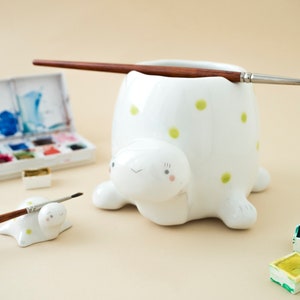 Puede incluir: Portacepillos de cerámica blanca con forma de tortuga con lunares verdes. Dos pinceles están en el portacepillos y una paleta de acuarela está en el fondo.