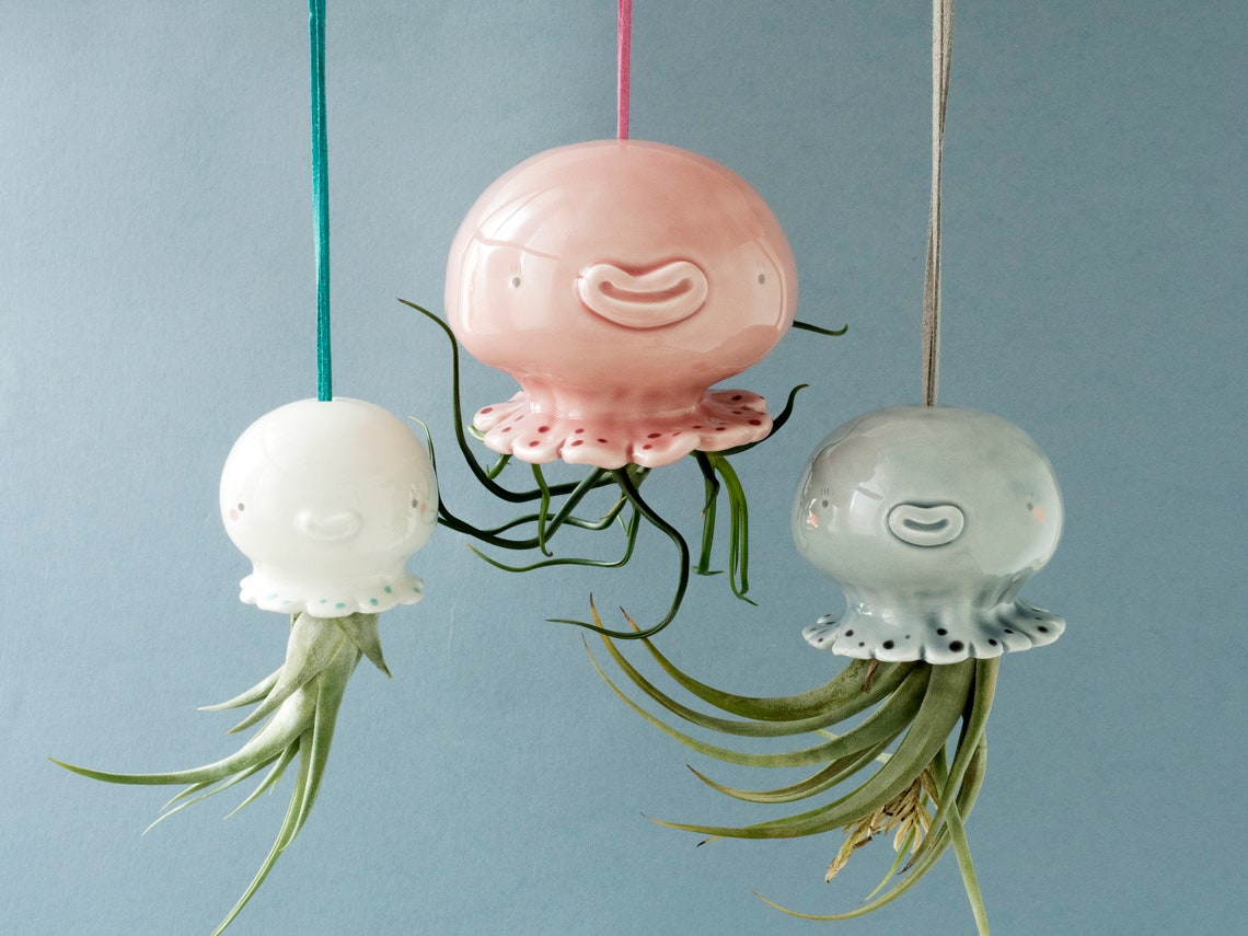 10 Fun Wall Hanging Planters To Bring Nature Indoor 4 May include: Three ceramic hanging planters shaped like jellyfish with air plants growing out of them. The jellyfish are white, pink, and light blue with black dots and painted faces. The planters are hanging from strings.
