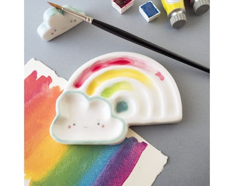 May include: A white ceramic rainbow-shaped paint palette with a cloud-shaped well. The palette has six wells for paint and a rainbow design painted on the surface. The cloud well has a cute face with eyes and a smile.