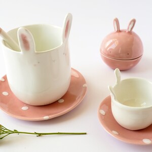 May include: A set of pink and white bunny-shaped ceramic teacups and saucers. The teacups have bunny ears and faces, and the saucers have pink polka dots.