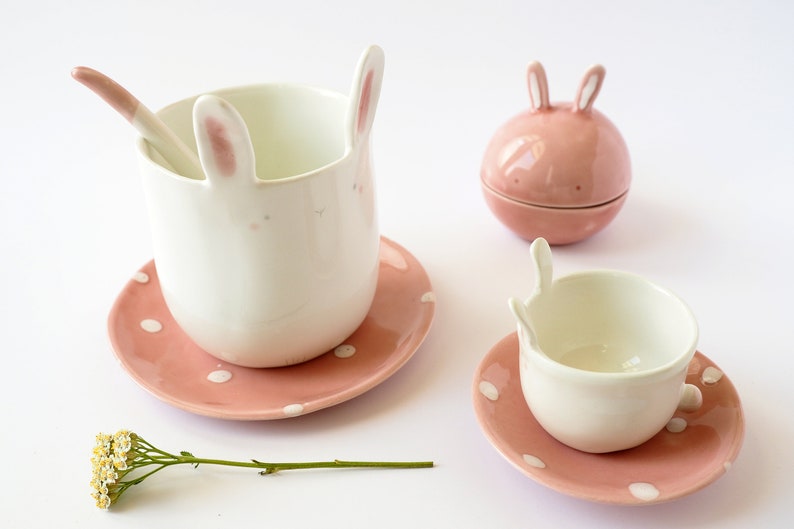 May include: A set of pink and white bunny-shaped ceramic teacups and saucers. The teacups have bunny ears and faces, and the saucers have pink polka dots.
