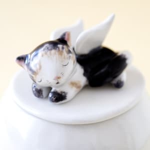 May include: White ceramic jar with a lid featuring a sleeping cat figurine with wings. The cat is painted with black, white, and brown markings.