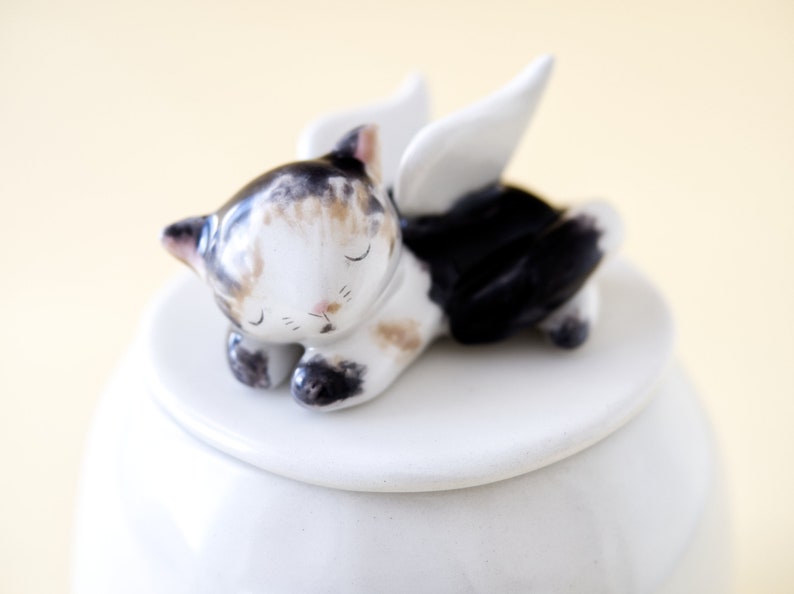May include: White ceramic jar with a lid featuring a sleeping cat figurine with wings. The cat is painted with black, white, and brown markings.