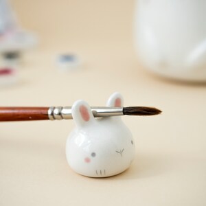 May include: White ceramic bunny brush holder with pink ears and a paintbrush resting on top.