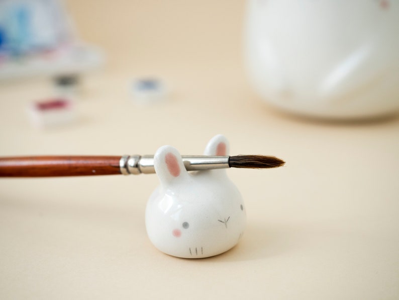 May include: White ceramic bunny brush holder with pink ears and a paintbrush resting on top.