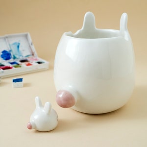 May include: White ceramic planter shaped like a rabbit with a pink nose and ears. The planter is sitting on a light brown surface.
