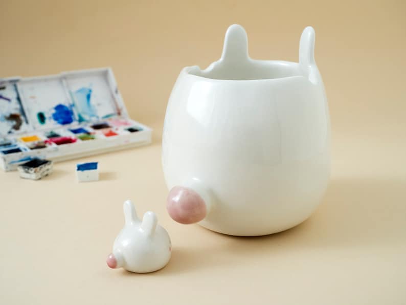 May include: White ceramic planter shaped like a rabbit with a pink nose and ears. The planter is sitting on a light brown surface.
