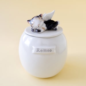 May include: White ceramic pet urn with a sleeping cat figurine on the lid. The cat has black, white, and brown fur and angel wings. The urn has the name "Romeo" written on it.