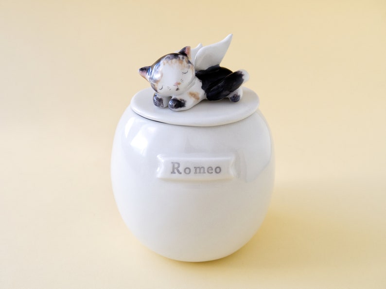 May include: White ceramic pet urn with a sleeping cat figurine on the lid. The cat has black, white, and brown fur and angel wings. The urn has the name "Romeo" written on it.