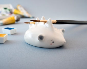 May include: White ceramic hedgehog brush holder with two paintbrushes resting in the spines. The hedgehog has a pink nose and black eyes.