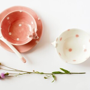 May include: Two pink and white polka dot ceramic bowls, one with a saucer and a spoon, and the other with a handle shaped like a rabbit.