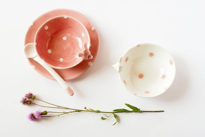 May include: Two pink and white polka dot ceramic bowls, one with a saucer and a spoon, and the other with a handle shaped like a rabbit.