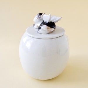 May include: White ceramic jar with a lid featuring a small, white, black, and brown ceramic figurine of a cat with wings.