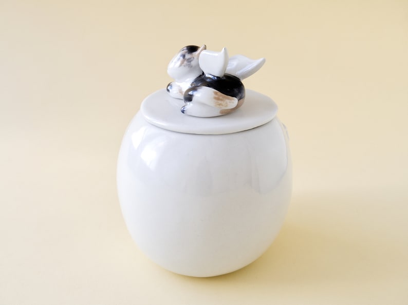 May include: White ceramic jar with a lid featuring a small, white, black, and brown ceramic figurine of a cat with wings.