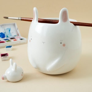 May include: A white ceramic bunny-shaped brush holder with a small bunny figurine next to it. The brush holder has pink ears and a painted face. A paintbrush is resting on the top of the holder.