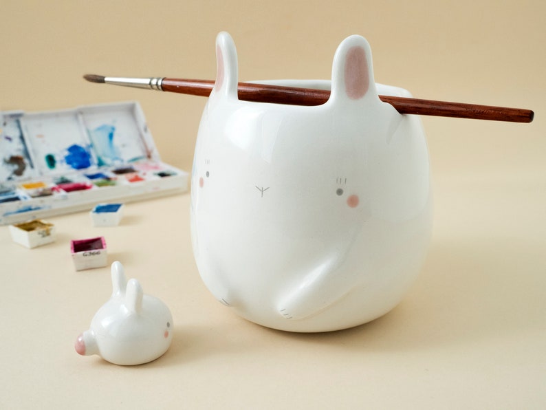 May include: A white ceramic bunny-shaped brush holder with a small bunny figurine next to it. The brush holder has pink ears and a painted face. A paintbrush is resting on the top of the holder.