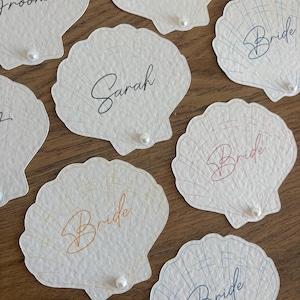 May include: Off-white seashell-shaped place cards with delicate line detailing. Some cards feature the word "Bride" in various colors, while others have names like "Sarah." Each card has a small pearl accent.