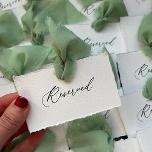 May include: A stack of white cards with the word "Reserved" written in black calligraphy. Each card is tied with a sage green ribbon.